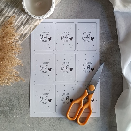 Minimalistische DIY-Neujahrskarte mit Wunderkerze und dem Text „Ein kleines Licht für 2024“, dekoriert mit Kerze, Trockenblumen und Stoff auf hellgrauem Untergrund.