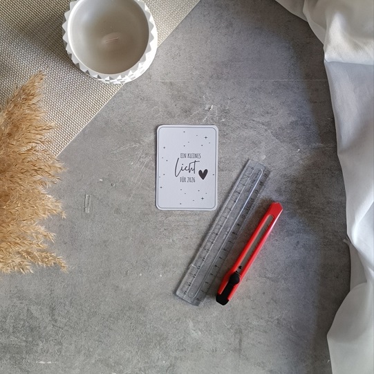 Minimalistische DIY-Neujahrskarte mit Wunderkerze und dem Text „Ein kleines Licht für 2024“, dekoriert mit Kerze, Trockenblumen und Stoff auf hellgrauem Untergrund.