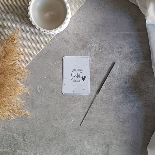 Minimalistische DIY-Neujahrskarte mit Wunderkerze und dem Text „Ein kleines Licht für 2024“, dekoriert mit Kerze, Trockenblumen und Stoff auf hellgrauem Untergrund.