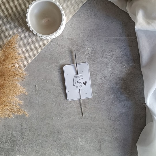 Minimalistische DIY-Neujahrskarte mit Wunderkerze und dem Text „Ein kleines Licht für 2024“, dekoriert mit Kerze, Trockenblumen und Stoff auf hellgrauem Untergrund.