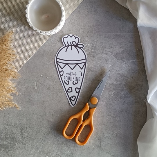 DIY-Karte in Schultütenform mit rotem Bleistift und dem Schriftzug „Endlich Schulkind“, dekoriert mit Kerze, Pampasgras und Stoff auf grauem Untergrund.